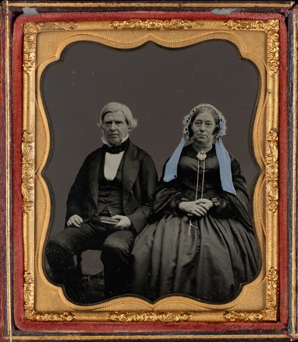The image shows a seated couple captured in a half-body portrait. The man on the left has short, gray hair, deep-set eyes, and a serious expression. He wears a black suit with a vest and a bow tie. The woman on the right has medium-length gray hair with a decorative headpiece and a composed look. She wears a dark dress with a light blue shawl, a necklace, and holds her hands together in her lap. The background is a simple dark backdrop. The photograph is in an ornate, gold-trimmed frame with intricate designs, set against a red velvet mat.