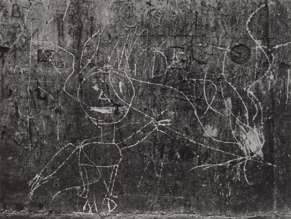 This image features white etched lines forming abstract shapes on a rough, textured surface. The central design resembles a simplistic human figure, with a circular head, facial features, and an angular body with stick-like limbs. Swirling lines extend outward from the figure's head, resembling tangled hair or flames. The background is filled with additional engraved marks, adding to the complexity and texture of the scene.