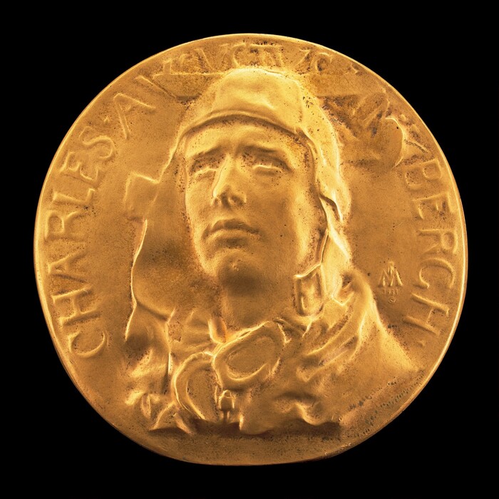 The sculpture is a circular, bas-relief medallion featuring the head and shoulders of a man in aviator attire. The man is wearing a helmet and goggles. The surface of the medallion is engraved with the name "CHARLES A. LINDBERGH." The sculpture is gold in color, made of a metallic substance, possibly bronze or a similar metal with a gilded finish. The man's face is depicted realistically with a determined expression, looking forward and slightly upward. The detailing in the sculpture captures aspects of the aviator's gear. On the right side, there is a small emblem or initials to the artist. The sculpture is presented against a black background.