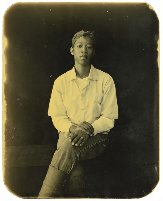 The image shows a person in a seated position with their body visible from the waist up. They are sitting with one leg crossed over the other, and their hands are placed on their knee. The individual has short hair covered by a patterned headscarf, tied at the front. They are wearing a light-colored, long-sleeved shirt over a t-shirt, and denim jeans. On one of their fingers, they wear a ring. In the background, there is a plain, dark surface that contrasts with the lighter tones of their clothing. The portrait has rounded corners on the photograph.