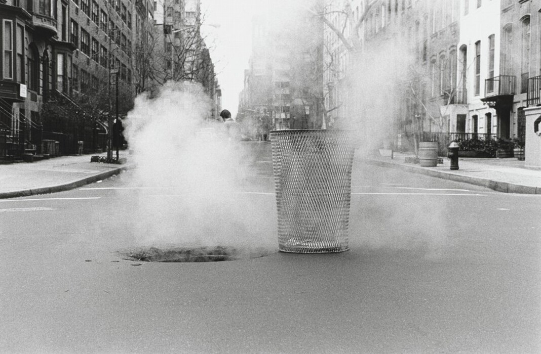 The image shows a black and white photograph of a wire mesh trash can on a city street. Steam is rising from a manhole or vent beside the trash can. The street is deserted, with buildings on either side leading to a vanishing point in the distance.