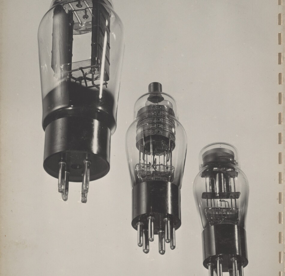 Three lightbulb-like objects with sockets at their bases are arranged in a diagonal row against a blank background in this black and white photograph. All three have cage-like components with wiring inside the bulbs but the designs differ. The largest bulb, to the left, has four prongs on the base, and the rectangular component inside fills most of the bulb. The other two are slightly smaller, and each has six prongs on the base. The cage-like component is round inside the middle bulb and a dark tip protrudes from the top of the bulb. The third has a square-shaped cage and a flat top. Vertical slits presumably from a previous binding line the right side of the white page.