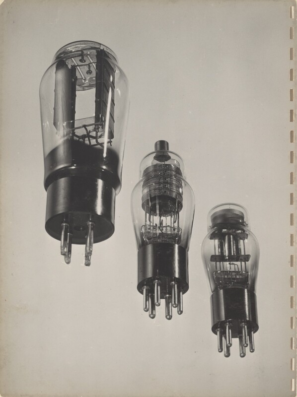 Three lightbulb-like objects with sockets at their bases are arranged in a diagonal row against a blank background in this black and white photograph. All three have cage-like components with wiring inside the bulbs but the designs differ. The largest bulb, to the left, has four prongs on the base, and the rectangular component inside fills most of the bulb. The other two are slightly smaller, and each has six prongs on the base. The cage-like component is round inside the middle bulb and a dark tip protrudes from the top of the bulb. The third has a square-shaped cage and a flat top. Vertical slits presumably from a previous binding line the right side of the white page.