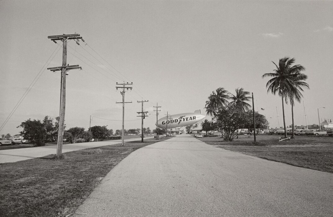 This grayscale photograph depicts a slightly curved road leading into the distance. On the left, several utility poles with wires stretch toward the horizon. A blimp with the word "GOODYEAR" is prominently visible in the center, hovering above the road. To the right, palm trees and parked cars line the road. The sky is clear with few clouds. The horizon is positioned in the center of the frame, emphasizing the open sky and creating a sense of space.