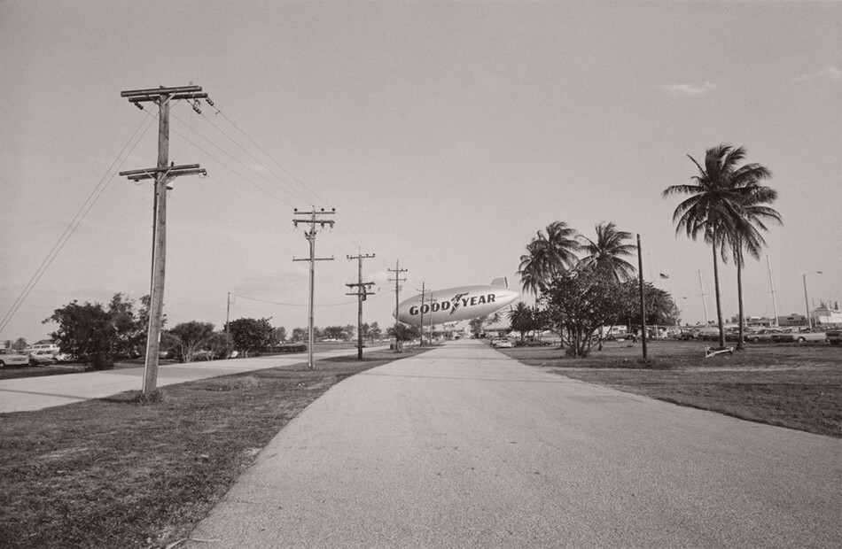 This grayscale photograph depicts a slightly curved road leading into the distance. On the left, several utility poles with wires stretch toward the horizon. A blimp with the word "GOODYEAR" is prominently visible in the center, hovering above the road. To the right, palm trees and parked cars line the road. The sky is clear with few clouds. The horizon is positioned in the center of the frame, emphasizing the open sky and creating a sense of space.