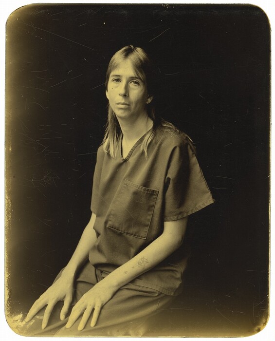 The image shows a person seated from the waist up with hands resting on their lap. The individual has long, straight hair parted in the middle, distinct facial features, calm expression, and eyes looking at the camera. They wear a short-sleeved outfit resembling a scrub top with a chest pocket, and a tattoo on their left forearm. A chain necklace is around their neck. The background is plain and dark.