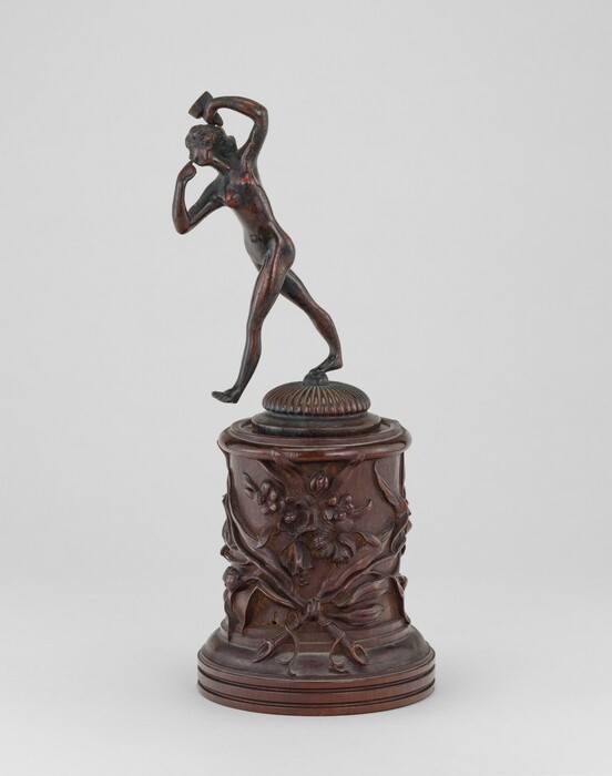 The sculpture depicts a nude figure of a slender and athletic individual in a dynamic pose, seemingly in motion. The figure is looking straight ahead with one arm raised overhead and the other bent. The sculpture is made from a material that appears to be a dark, polished bronze. The figure stands atop a carved pedestal that is adorned with floral motifs and ribbon-like designs. The pedestal is crafted from a wood-like material or has a wooden finish, with a rich, brown color. The sculpture stands as a testament to classical sculptural artistry.