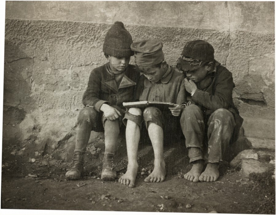 This is a photograph of three children seated closely together outdoors. The children are looking at a paper held by the child in the center. They are shoeless and appear to be engaged in reading or looking at the paper.