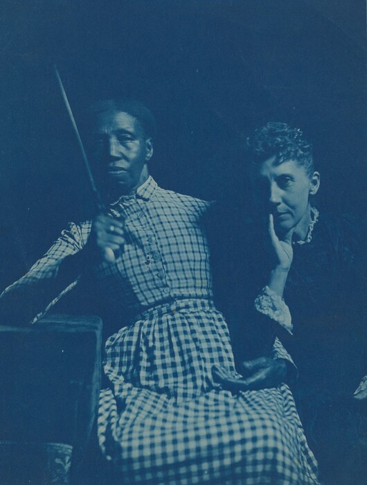 The image shows two people from the waist up. The person on the left is sitting slightly forward and is holding a large, thin, cylindrical object. They have short, dark hair and are wearing a high-necked, long-sleeved, checkered dress with white and dark checks. Their facial expression is neutral, with their eyes looking straight ahead. The person on the right is sitting close to the first person, with their hand resting on the side of their face, and they have light curly hair. They are wearing a dark garment with a light-colored collar. The background is dark.