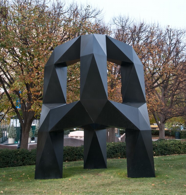 This free-standing sculpture is a three-footed, black metal arch with flat, faceted surfaces along its chunky legs. On the top half, three arms curve up and in to meet at the center. The arms are offset from the three legs and are connected by bands across the middle. The sculpture stands on a grassy park lawn and is nearly as tall as the mature trees behind it. Sunlight gleams on the matte surfaces so some look lighter gray and others are flat black.