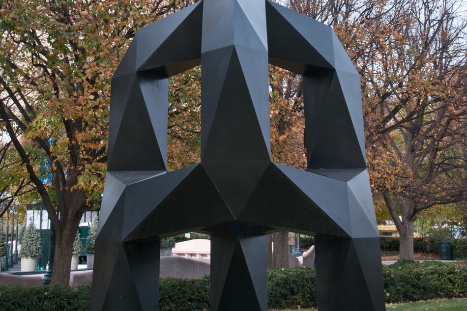 This free-standing sculpture is a three-footed, black metal arch with flat, faceted surfaces along its chunky legs. On the top half, three arms curve up and in to meet at the center. The arms are offset from the three legs and are connected by bands across the middle. The sculpture stands on a grassy park lawn and is nearly as tall as the mature trees behind it. Sunlight gleams on the matte surfaces so some look lighter gray and others are flat black.