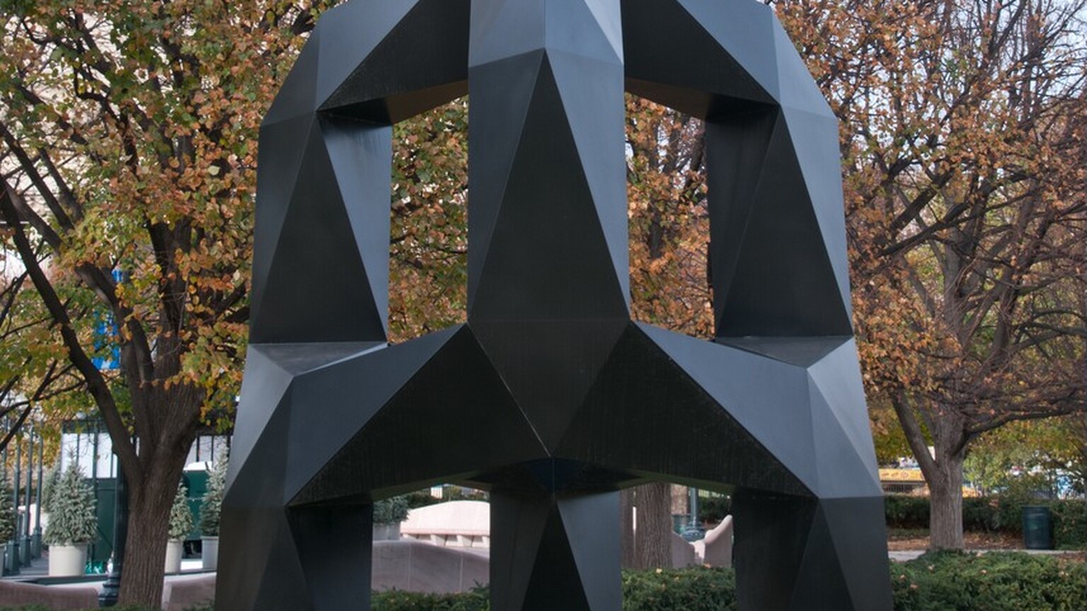 This free-standing sculpture is a three-footed, black metal arch with flat, faceted surfaces along its chunky legs. On the top half, three arms curve up and in to meet at the center. The arms are offset from the three legs and are connected by bands across the middle. The sculpture stands on a grassy park lawn and is nearly as tall as the mature trees behind it. Sunlight gleams on the matte surfaces so some look lighter gray and others are flat black.