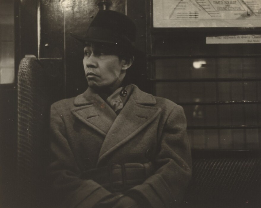 The image shows a man sitting with his upper body depicted. He is turned slightly to his right, looking off to the side. He has short hair covered by a dark fedora hat. The man appears to be of Asian descent and wears a large, buttoned-up overcoat with a belt. A portion of a shirt collar is visible underneath. The background shows a wooden wall with a grid-like structure and a partial poster on the right. The setting resembles an interior with upholstered seating, suggesting it might be a train or subway car. There are no visible pieces of jewelry or objects in his hands, and the lighting is dim, creating a moody atmosphere.