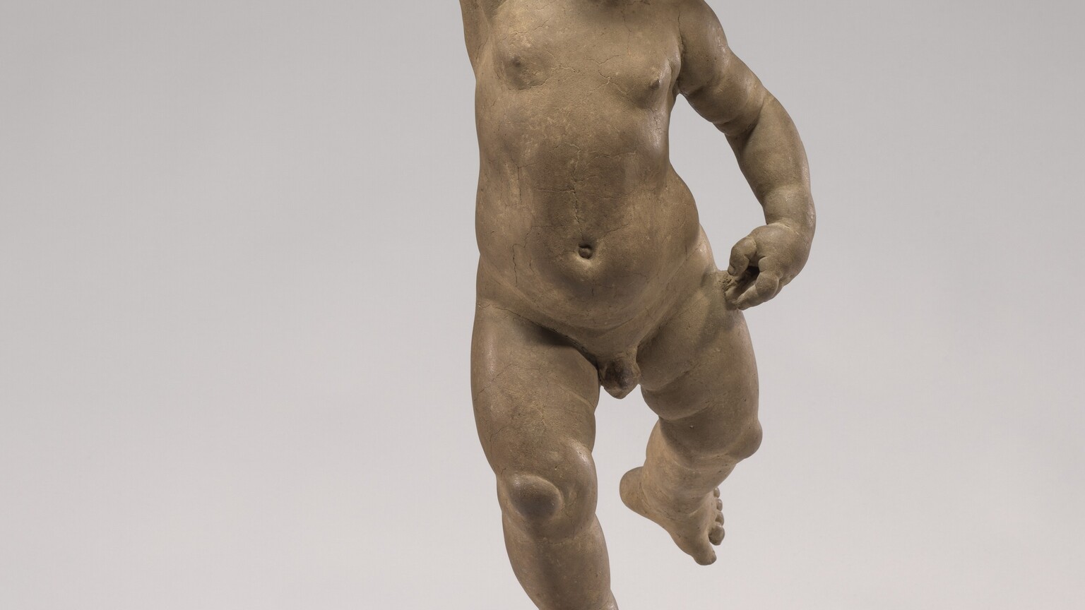 Sculpted in clay, a toddler balances his plump body on his right foot as he raises his left leg and right arm. In ths photograph, his body is squared to us. His left hand, on our right, rests near his hip. Gazing up and to our left, his cheeks are puffed in blowing through pursed lips, and his hair falls in soft ringlets. The fingers of his raised hand are folded into a loose fist. Tipped slightly forward onto his toes, he balances on a gold-half dome.
