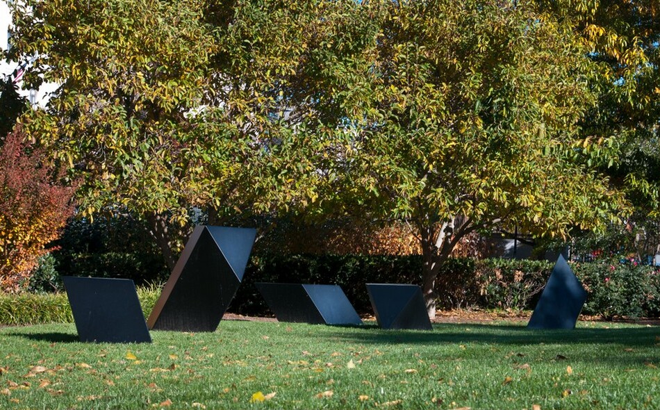 The sculpture features a row of geometric forms on a grassy field. Each piece is a parallelogram-like shape with several corners and flat sides, all made from a dark material and varying in size and angle. They are positioned in a park setting with trees and a well-maintained lawn.