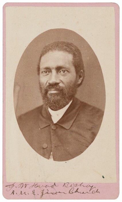 The image shows a man facing the camera in a seated position. He has distinct facial features with a full beard and mustache. The man wears a dark jacket with a high collar and a white shirt underneath. The background is plain and monochromatic. A card with handwritten text is placed below the image.
