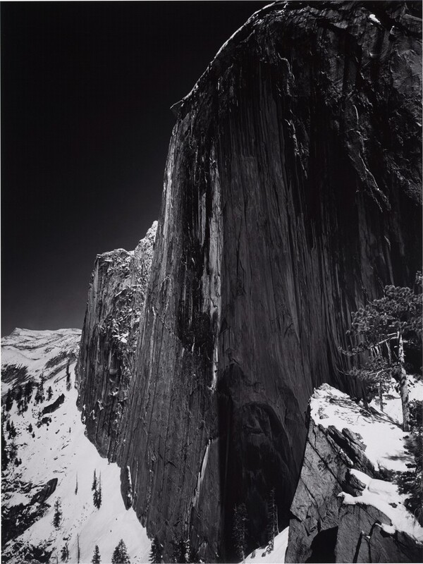 A tall, almost sheer rock formation soaring high above snow-capped rocky peaks nearly fills this vertical black and white photograph. The rock formation is rounded along the top and continues off the top right corner and right edge of the composition. A jagged ridge dips down like a narrow wing to our left, and tall trees on the mountain peaks are made minuscule by the formation, the surface of which is textured with long vertical streaks. The sky in the upper left corner of the photograph is steely gray behind a thin ridge of white snow along the curved top of the formation.