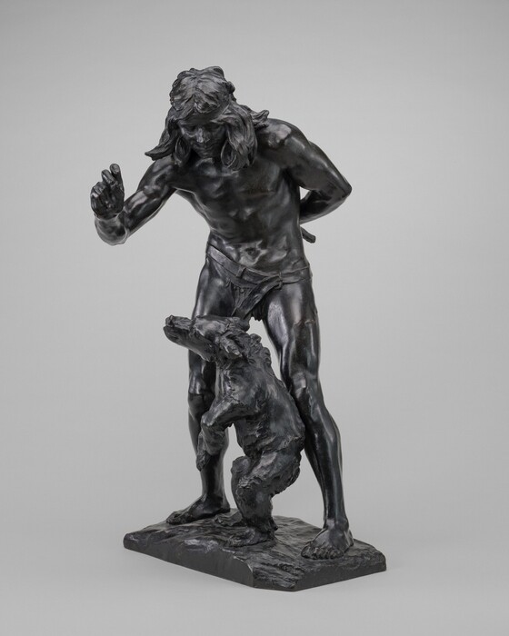The sculpture depicts a male figure with long hair in a dynamic pose. His left arm is bent with the fist raised, while his right arm is positioned behind his back. The figure is wearing a loincloth. A bear stands on its hind legs in front of him. The sculpture is made of a dark material, possibly bronze. The man's musculature and the bear's fur are detailed. The sculpture is presented on a textured rectangular base.