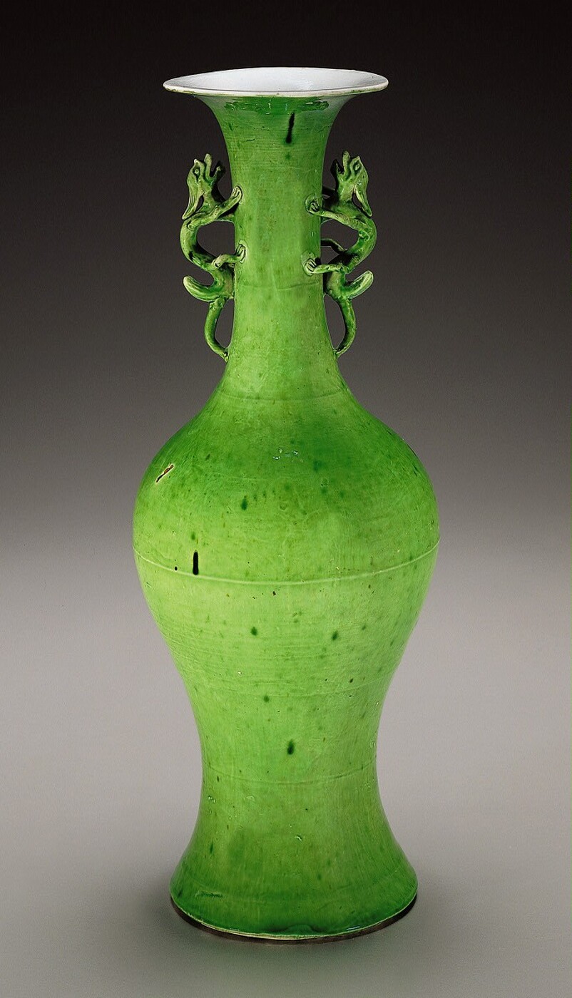 The sculpture is a decorative vase with two dragon-like handles on either side of its neck. The vase has a bulbous base that narrows into a slender neck, then flares out at the rim. It is made of ceramic with a green glaze, speckled with darker spots. The vase is upright with a symmetrical and balanced design.