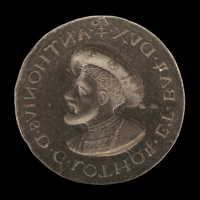 The sculpture features a man in profile facing left. He wears a hat with a diamond-shaped pattern and detailed textured clothing. The silver-colored metal sculpture has raised text along the outer edge. The piece is flat, round, and finely crafted.