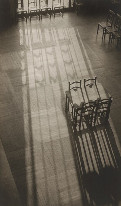Several chairs and a table are arranged on a wooden floor, with prominent shadows cast by the objects. The color palette consists of shades of gray due to the lighting. The table and a pair of chairs are positioned on the right side and slightly below the center of the image, with additional chairs lined up against windows near the top edge. The shadows of the window frames and furniture overlap on the floor.