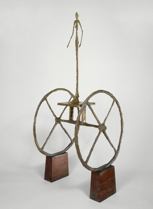 An abstracted, nude person stands on a platform perched on two wheels, all cast in bronze, which balances on wooden blocks in this freestanding sculpture. The elongated, unnaturally thin woman stands with her arms slightly bent and held out to her sides. She has low-set breasts, and her blocky, oversized feet stand on a metal platform supported on two vertical strips of bronze that perch on the axle. The wheels are oversized with four spokes each arranged to look like Xs. The surface of the woman and wheels are rough and knubby, and all of the metal is colored gold. The wood blocks stand upright with channels cut into their upper surfaces to hold the wheels. The sculpture is angled to our left in this photograph, and it is shown against a pale gray background.