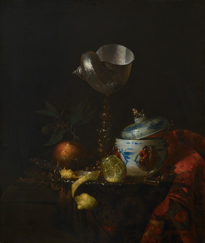 A tall goblet made with a spiraling nautilus shell on a gold and silver stem, a blue and white lidded, porcelain bowl, an orange, a cut lemon, and a bunched up, woven rug are gathered on a tabletop in front of a black background in this vertical still life painting. Light falling on the objects from our left contrasts sharply with the deep shadow beyond, which makes some details difficult to make out. The objects might be gathered on a silver tray. At the center of the composition, the flagon is made with a shimmering, ivory-colored nautilus shell with its wide opening facing up. The stem is made of gold bands coiling around a silver figurine, which is nearly lost in shadow. To our right, a white porcelain bowl has a rounded bottom and tall sides. It is painted with blue designs around a raised relief showing two people in crimson-red and harvest-gold garments. A utensil sits in the bowl to our right, and the lid, which has a finial in the shape of an animal, rests angled upward, partially in the bowl and partially on the stem of that utensil. A translucent, cylindrical knife handle protrudes toward us, angled to our right, in front of the bowl. A rug with a geometric pattern of rust red, tan, and pine green is bunched next to the bowl, along the right side of the painting. To our left of the nautilus cup, an orange with a stem and green leaves sits next to a bright, glistening lemon at the foot of the cup. Between the two pieces of fruit, the vivid yellow lemon rind has been peeled into a long, curling strip that curves off the front edge of the table.