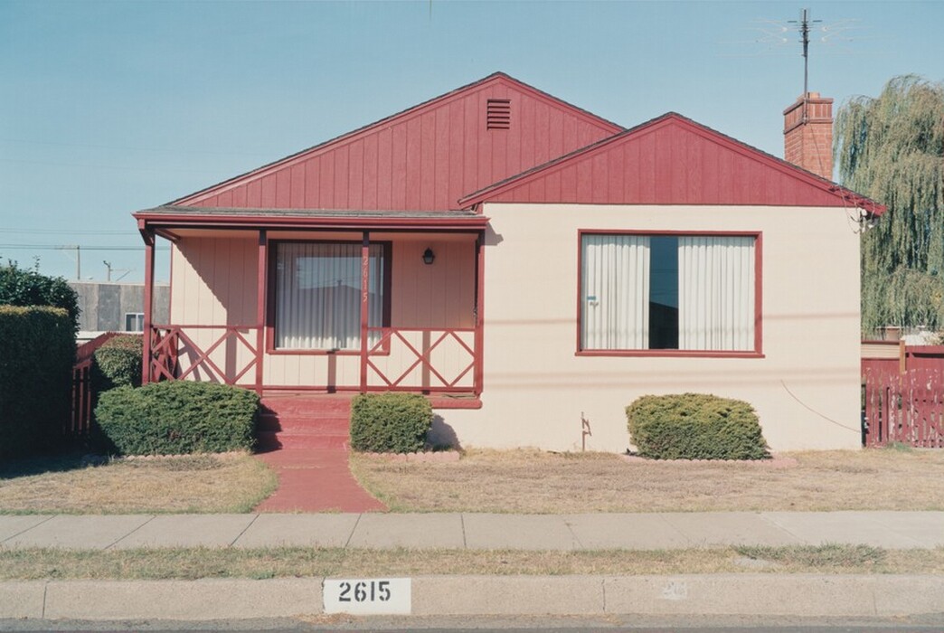 The image is a straightforward, head-on view of a single-story house. The house is painted with peach-colored walls and a deep red trim. The composition is symmetrical, with the house number "2615" visible on the curb and porch post. The front yard has short hedges and a red pathway leading to the front door. The image is framed by a clear blue sky, a television antenna on the right side, and a tree behind the house.
