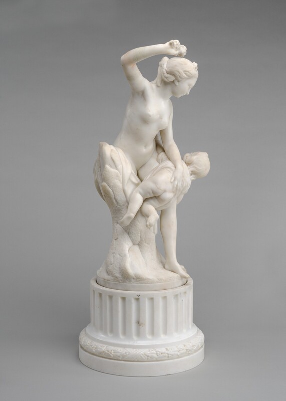 A nude woman stands and braces a child over her knee in this freestanding white marble sculpture. In this photograph, the woman’s body is angled to our right as she raises her right arm, which is closer to us. She grips a bouquet of flowers in that hand, her elbow bent. She has a delicate profile, and her wavy hair is pulled back and plaited under a ribbon and flowers. Leaning to our left against a trunk-like support, the woman grips the child’s back, holding his garment up over his nude backside. His arms splay wide, and he kicks one foot. The sculpture is on a fluted, round base with stepped molding at the bottom, and the background is pale gray.