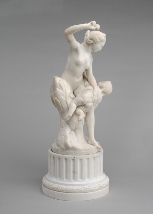 A nude woman stands and braces a child over her knee in this freestanding white marble sculpture. In this photograph, the woman’s body is angled to our right as she raises her right arm, which is closer to us. She grips a bouquet of flowers in that hand, her elbow bent. She has a delicate profile, and her wavy hair is pulled back and plaited under a ribbon and flowers. Leaning to our left against a trunk-like support, the woman grips the child’s back, holding his garment up over his nude backside. His arms splay wide, and he kicks one foot. The sculpture is on a fluted, round base with stepped molding at the bottom, and the background is pale gray.