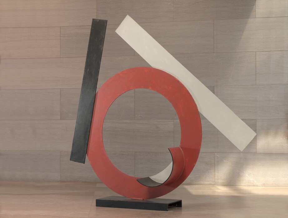 Made with sheets of painted steel, a red ring stands upright on a narrow black base in this freestanding sculpture. A black strip and a white strip are affixed to the edges of the ring. An inward-facing cup-shaped lip at the bottom right side of the circle is lined with black along the inner edge and painted white where it encloses space on the otherwise red ring. The black rectangular strip, which would be about the same height as the ring if stood alongside it, is affixed to the left center of the face of the ring almost vertically. The longer white strip is affixed to the back side of the ring and angles from near the top of the black strip down and to the right. The sculpture is photographed near a wall clad with pale pinkish-gray marble and a similarly colored floor.