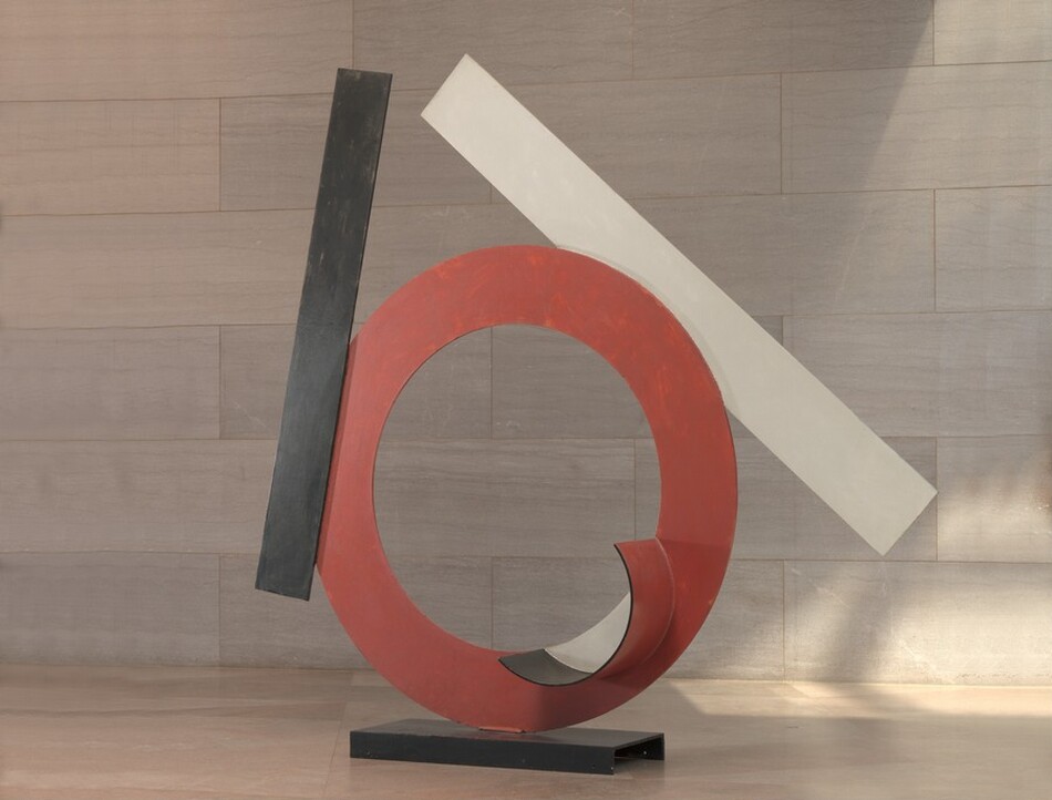 Made with sheets of painted steel, a red ring stands upright on a narrow black base in this freestanding sculpture. A black strip and a white strip are affixed to the edges of the ring. An inward-facing cup-shaped lip at the bottom right side of the circle is lined with black along the inner edge and painted white where it encloses space on the otherwise red ring. The black rectangular strip, which would be about the same height as the ring if stood alongside it, is affixed to the left center of the face of the ring almost vertically. The longer white strip is affixed to the back side of the ring and angles from near the top of the black strip down and to the right. The sculpture is photographed near a wall clad with pale pinkish-gray marble and a similarly colored floor.