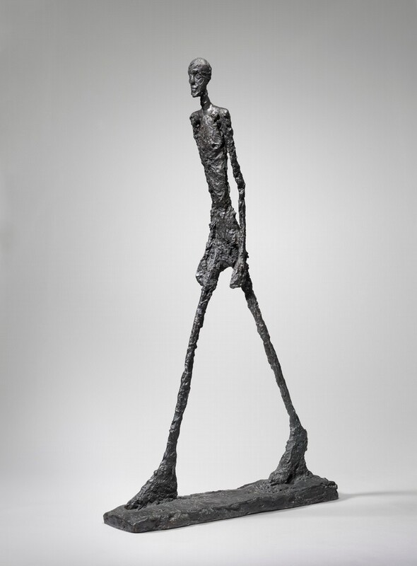 An abstracted nude man strides forward on long, spindly legs with his arms by his sides in this free-standing bronze sculpture. The man’s limbs are unnaturally thin and elongated with oversized hands and feet. The surface of the sculpture and the narrow, rectangular base is bumpy and rough. His body is angled to our right against a light gray background in this photograph.
