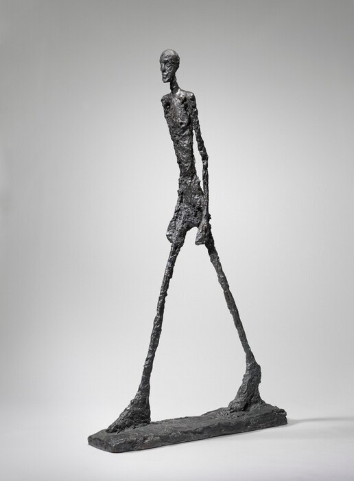 An abstracted nude man strides forward on long, spindly legs with his arms by his sides in this free-standing bronze sculpture. The man’s limbs are unnaturally thin and elongated with oversized hands and feet. The surface of the sculpture and the narrow, rectangular base is bumpy and rough. His body is angled to our right against a light gray background in this photograph.