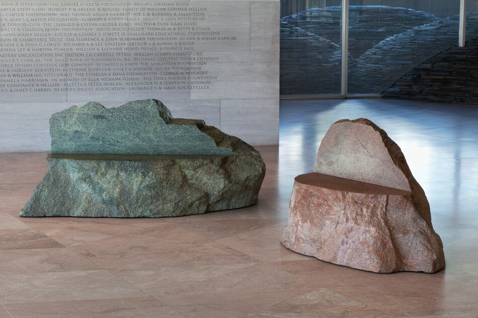 The image shows two stone sculptures resembling benches. The left sculpture is green with a textured surface. The right sculpture is reddish-brown, also with a rough surface. Both have flat top surfaces for seating. They are in an indoor space with a polished stone floor and a wall with engraved text, potentially names.