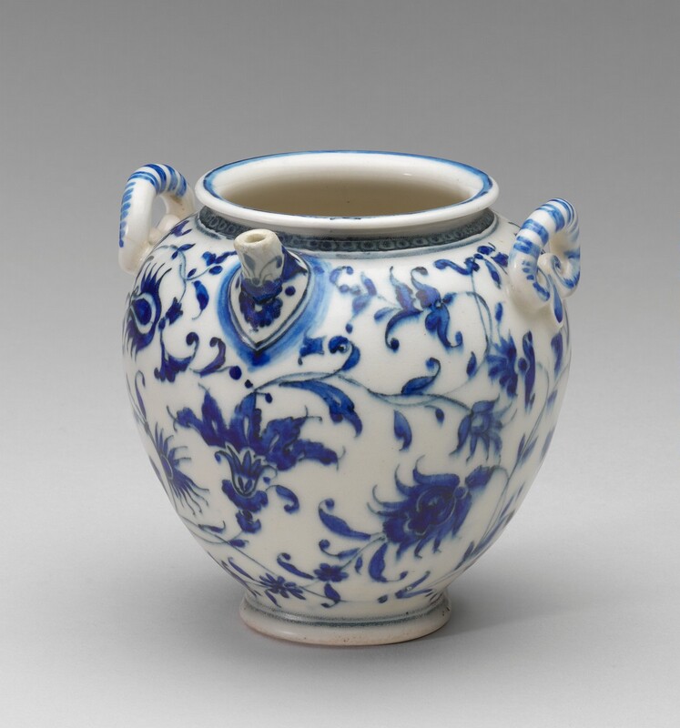 Blue floral patterns adorn this bulb-shaped white flask made of imitation porcelain. The round opening at the top of the flask has a low lip. The rounded vessel tapers from wide shoulders to a narrower flared foot at the bottom. There are two handles on either side of the top opening and a short spout near the rim directly between them. The rim is decorated with dots set within squares, and the handles are painted with blue stripes.