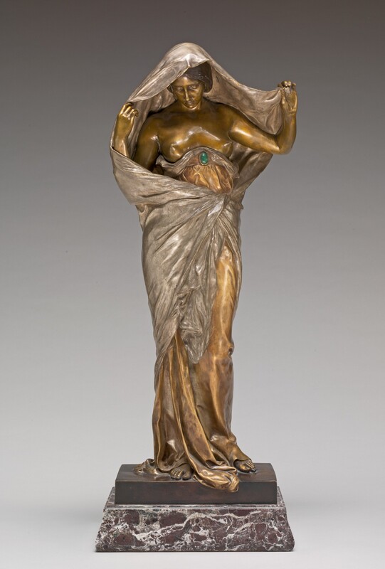 A woman wrapped in silver and gold robes lifts fabric to reveal her head and face in this freestanding cast bronze sculpture. The woman’s skin and a garment held in place with a belt are polished gold. The fabric that drapes over her head and wraps around her garment is silver, and her hair is dark brown. In this photograph, her body faces us, and she tips her head to look down and slightly to our left. She has a long nose, high cheekbones, a pointed chin, and her lips are closed. The belt has an oval green stone at the front, and it wraps tightly around her ribcage just under her bare breasts. The gold garment puddles around her bare feet, and a bit spills off the front edge of the brown, bronze square platform, which sits on a slightly wider marble base swirled with white and russet brown.