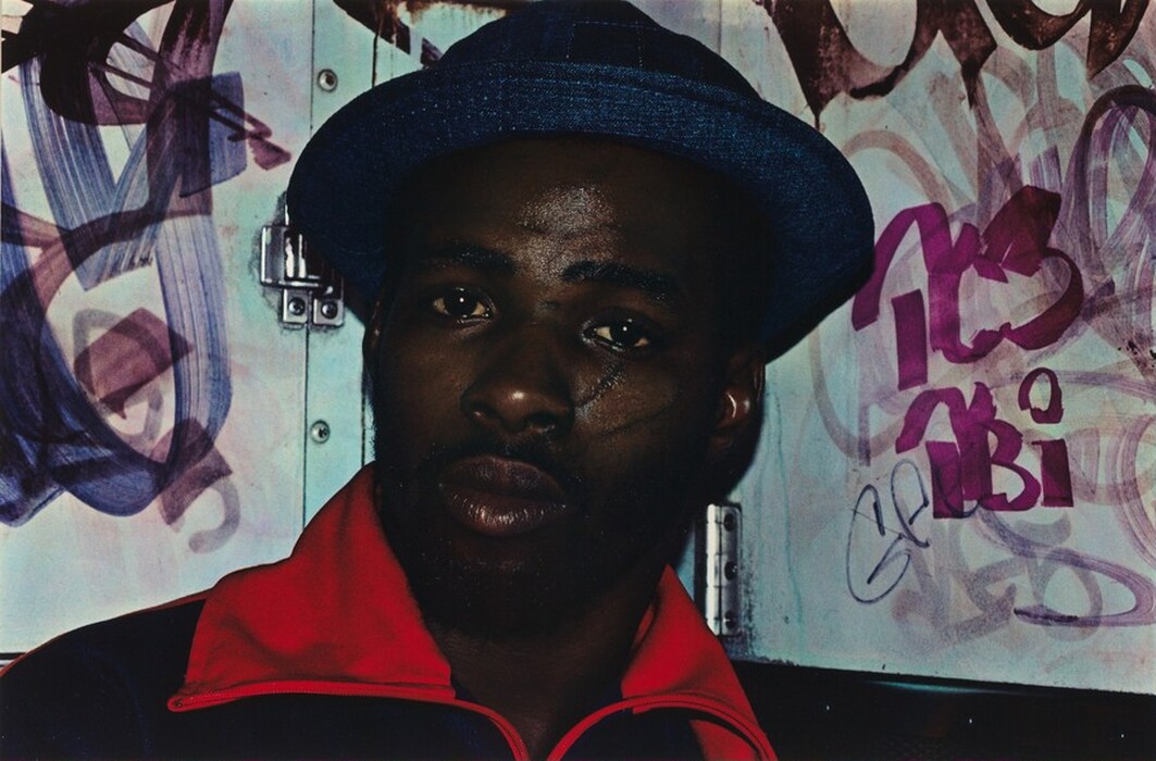 The image shows a man with dark skin and a beard wearing a red and dark-colored jacket and a blue textured hat. The man is facing the camera with a slightly turned head. In the background, there are various purple, pink, and grey graffiti on a wall and an industrial metal latch.