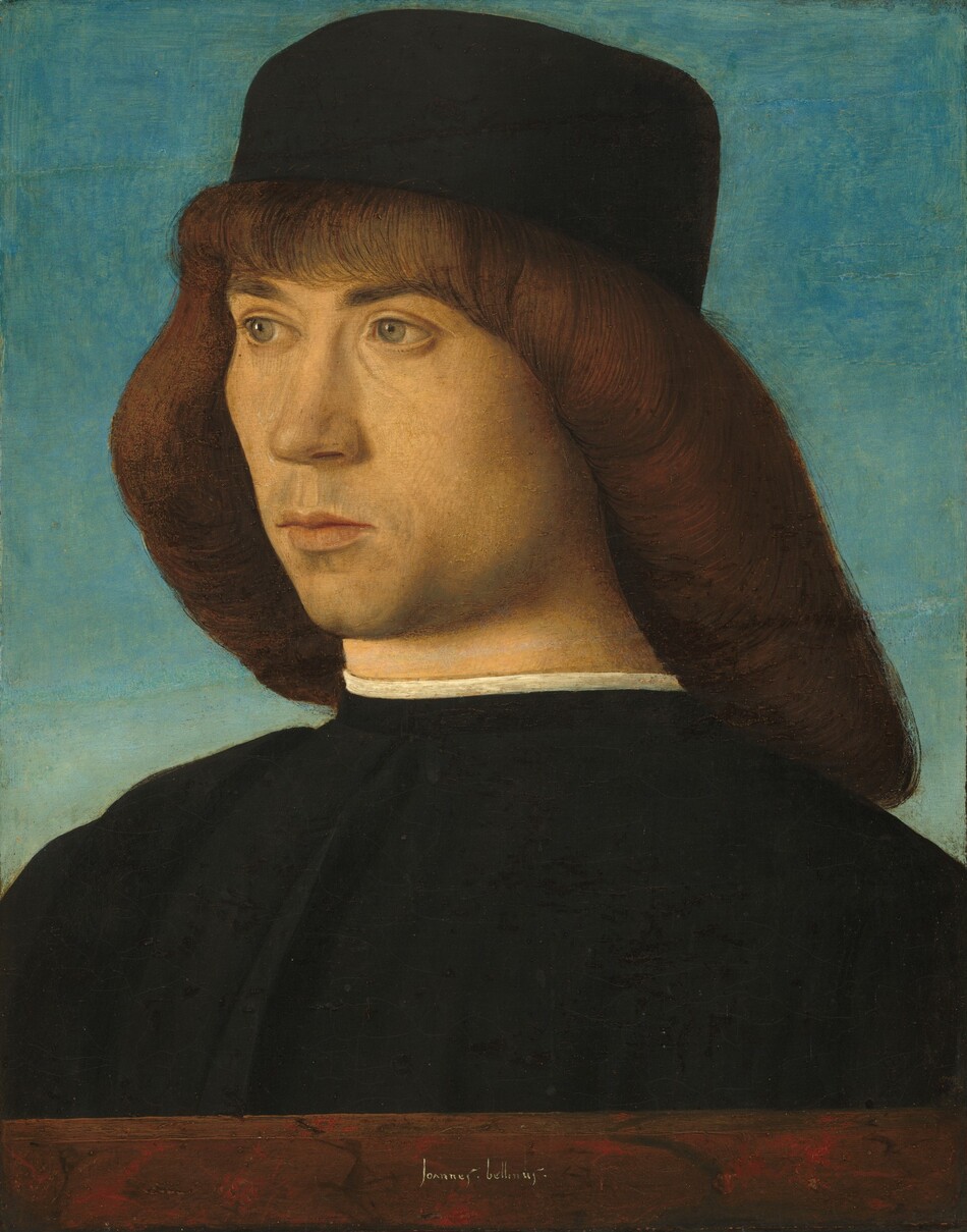 The head and shoulders of a pale-skinned, clean-shaven man fills this vertical portrait painting. The man’s shoulders are angled to our left, and he looks off in that direction with light green eyes. He has a straight nose, a long philtrum over pale pink lips, a cleft chin, and the suggestion of a five o’clock shadow. He has brown hair and bangs under a brimless, square-crowned cap. His hair curls under along the bottom to create a diagonal sweep from his bangs, over his ears, and to the back of his shoulders. The edge of a white undergarment is visible above the high neck of his black coat, which is pleated vertically down the front. The man is shown behind a ledge painted to look like red, brown, and black marble and the background is clear blue sky. The artist signed the front center of the ledge, “Joannes.bellinus.”