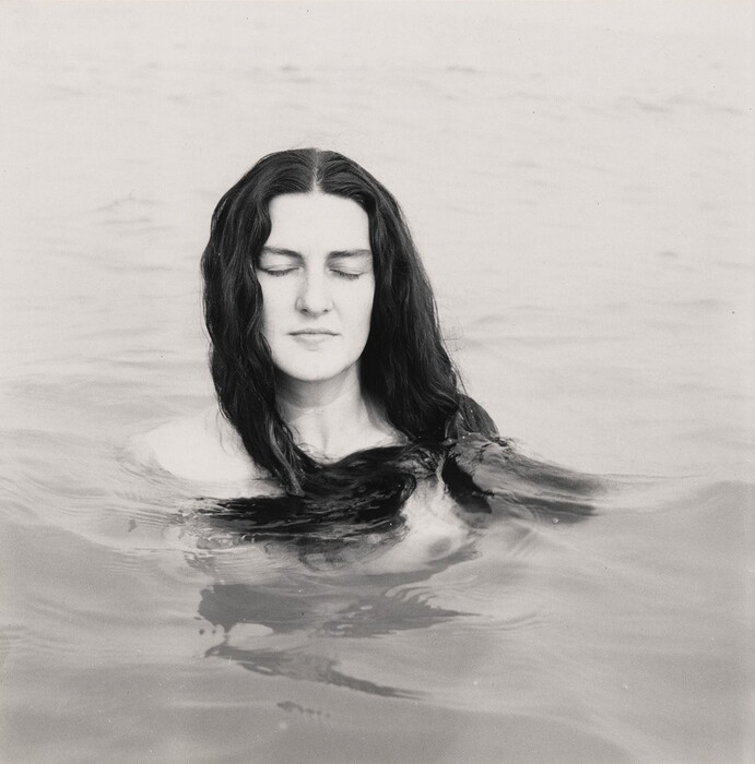 The image shows a person from the shoulders up, with the lower portion of their body submerged in water. The individual is in an upright position, facing forward with their eyes closed. They have long, dark hair that falls over their shoulders and into the water. Their facial features are relaxed with a calm expression, and the skin appears smooth and light-toned. No clothing is visible, as the person is mostly submerged, and there is no jewelry or objects being held. The background consists of gently rippling water, extending outwards, with no other elements visible beyond the natural water setting.