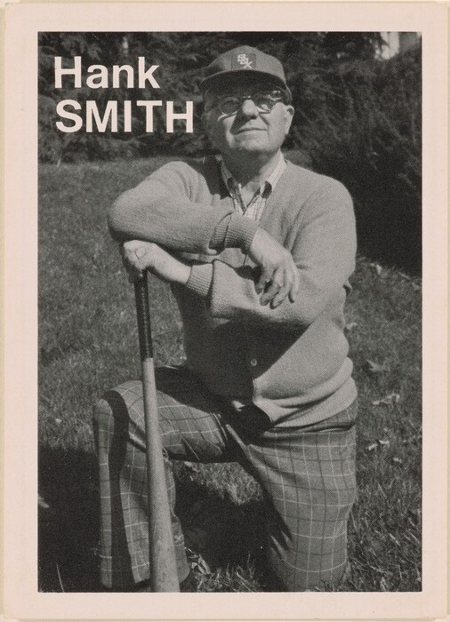 The image depicts a man in a kneeling position, with one knee on the ground and his hand leaning on a baseball bat. He is wearing glasses, a baseball cap, a sweater over a collared shirt, and checkered pants. The background shows a grassy area with trees or bushes. The words "Hank SMITH" are displayed to the left of the man.