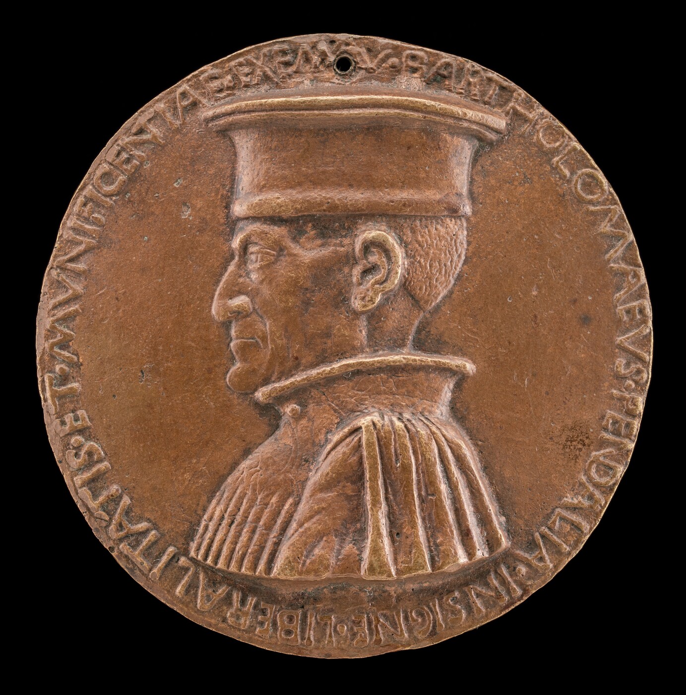 The sculpture is a circular medallion featuring a profile portrait of a man wearing a high-collared garment and a wide-brimmed hat that covers his head entirely. The subject is depicted in a side profile facing left, with distinct features such as a sharp nose, a pronounced chin, and a defined jawline. The material of the sculpture appears to be bronze, given its metallic, brownish hue, and slightly roughened texture. The surface of the medallion has inscriptions running along the perimeter, encircling the central portrait. The overall appearance gives the impression of a historical or commemorative item, with detailed craftsmanship evident in the relief work and the engraved lettering.