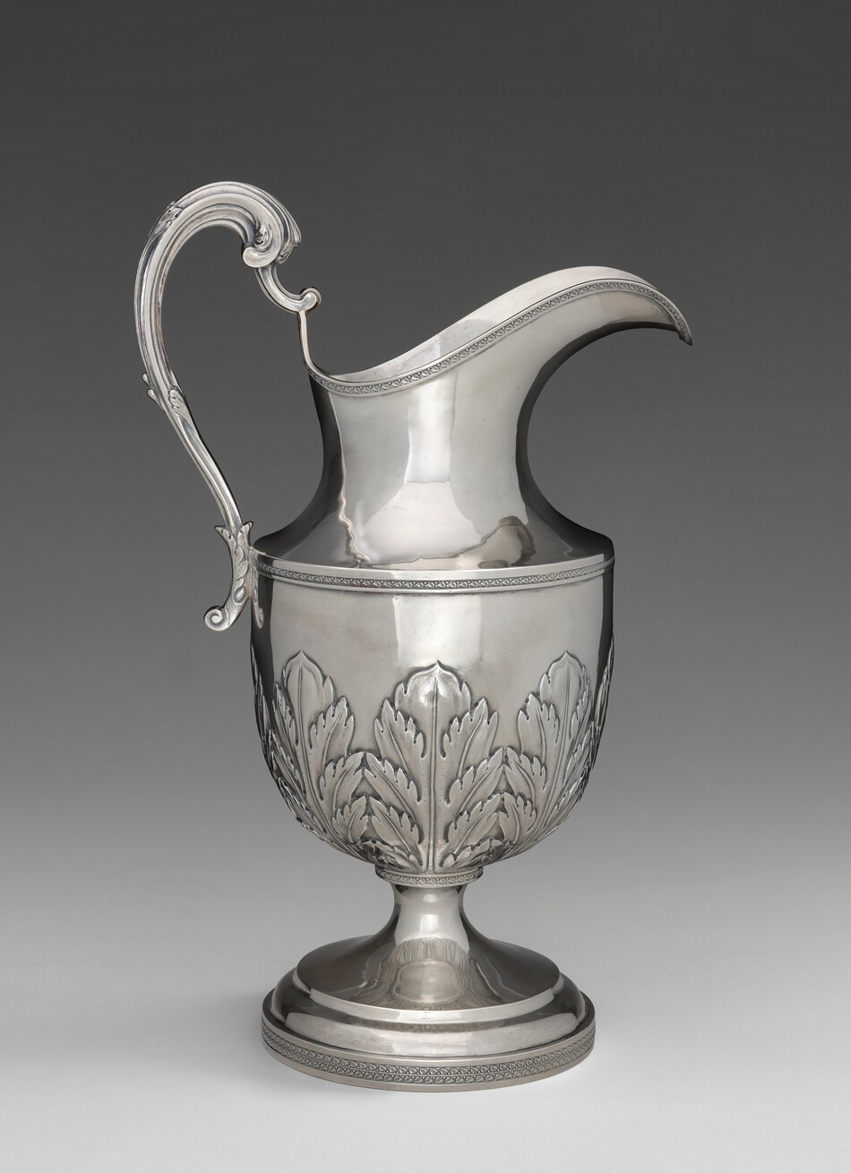 The image displays a silver-colored jug with a large body adorned with a leaf pattern and a curved handle. The color scheme is primarily metallic silver with highlights and reflections. The jug is centered in the image with the spout slightly angled to the right and the handle on the left side.