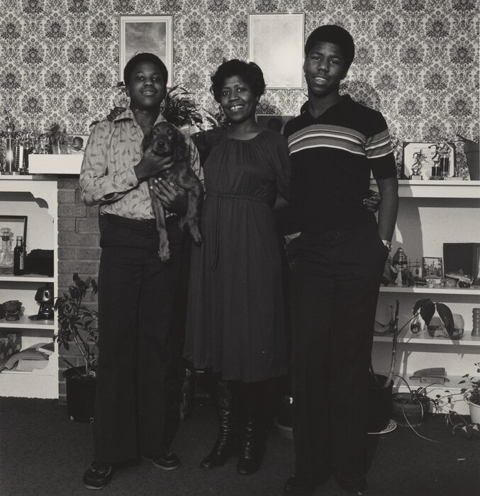 The image shows three people standing close together. On the left, a person stands with one arm around the middle person's shoulder, holding a dog in the other arm. The person is dressed in a patterned shirt and dark pants. The middle person, wearing a solid-colored dress, is positioned between the two others, smiling at the camera. The person on the right has short hair and is dressed in a striped top and dark pants. They all have short hair. They are standing in front of a patterned wallpaper and a decorated shelf filled with various items, including plants and trinkets. In the background, there is a fireplace and more shelves adorned with personal items, creating a homely setting.