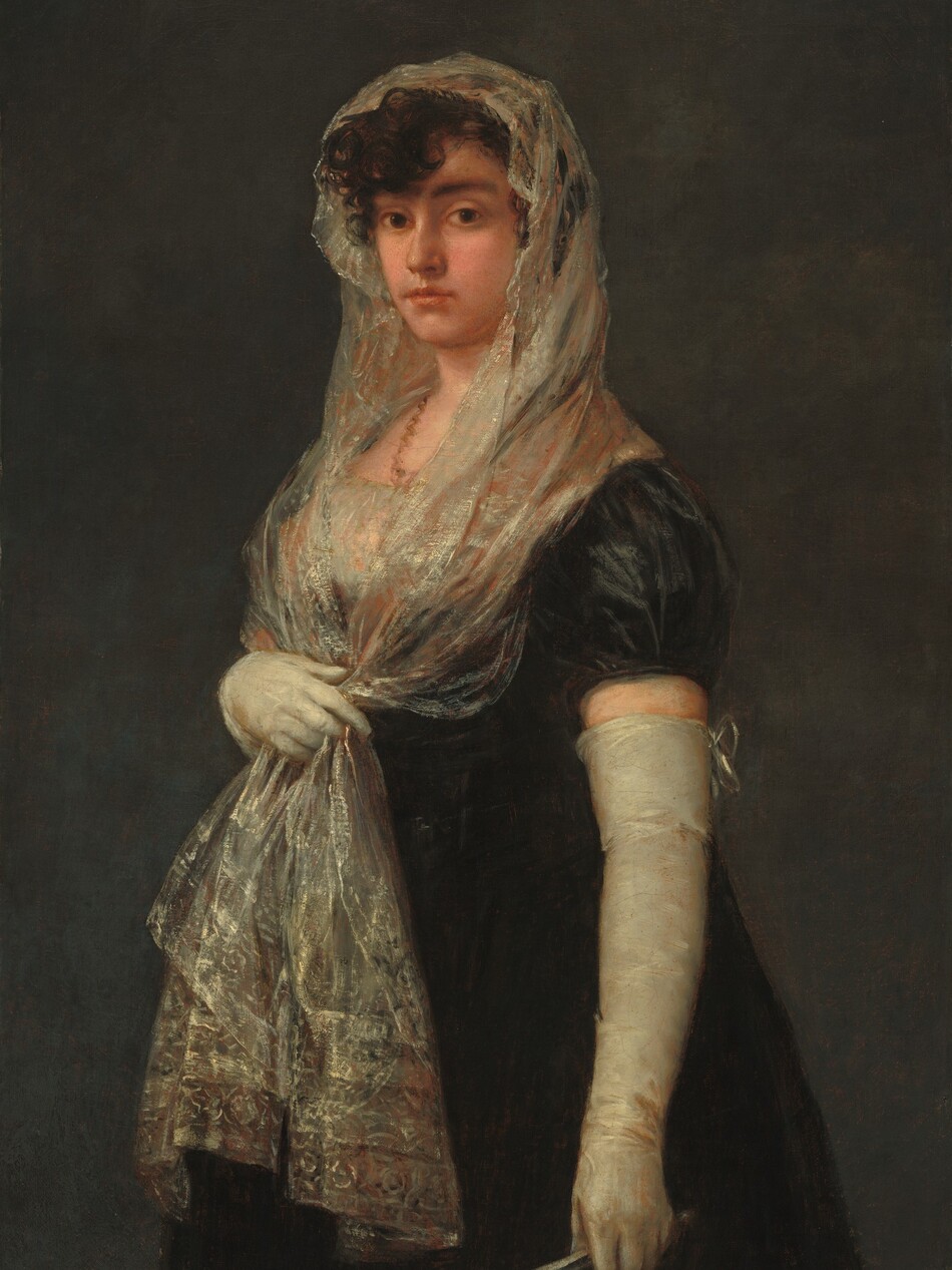 Shown from the knees up against a dark gray background, a young woman with peachy skin stands with her body angled to our left and she looks at us with brown eyes in this vertical portrait painting. She has dark, arched brows, a straight nose, and her full, coral-pink lips are closed. Her dark brown, curly hair is covered by a mantilla, an ivory-white, lacy scarf highlighted with pale gold and subtle, russet-red highlights. She holds the two long ends of the mantilla with her right hand, farther from us, below her bust. The ends of the scarf fall to mid-thigh over her long, ink-black dress. The bust of her dress is also white and shimmers, like the mantilla. Long, tight-fitting, off-white gloves nearly reach the short, puffed, cap sleeves of the dress. The back of the glove on her left arm, closer to her, is tied at the top with a bow, and she holds a silvery-white, closed fan in that hand. A brownish-coral colored necklace is painted as a squiggle of paint around her neck. Barely visible, the painter signed the painting in the lower left: “Goya.”