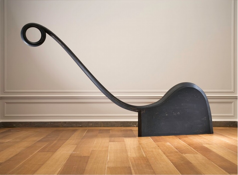 A long, thin arm ending in a round loop curves up and away from a snail shell-shaped panel in this freestanding, abstract sculpture. The arm is about twice as long as the panel, and it reaches to our left in this view. The sculpture is photographed in a room with a wood floor and a pale beige wall with molding.