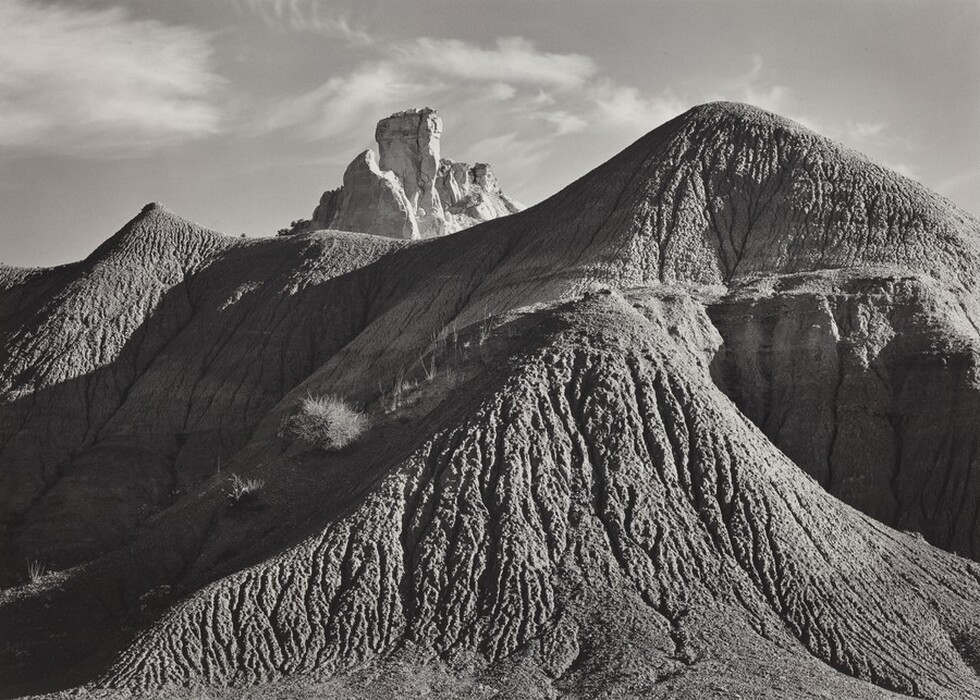 The image shows a landscape with undulating land formations leading towards a towering rock structure. The horizon is high in the composition, with a rock peak extending into the sky, under a sky with soft clouds. The texture of the earth is detailed, with erosion patterns. The color palette is monochromatic, emphasizing light and shadow contrasts. The play of highlights and shadows creates a dynamic visual effect, highlighting the rugged surface of the landscape.