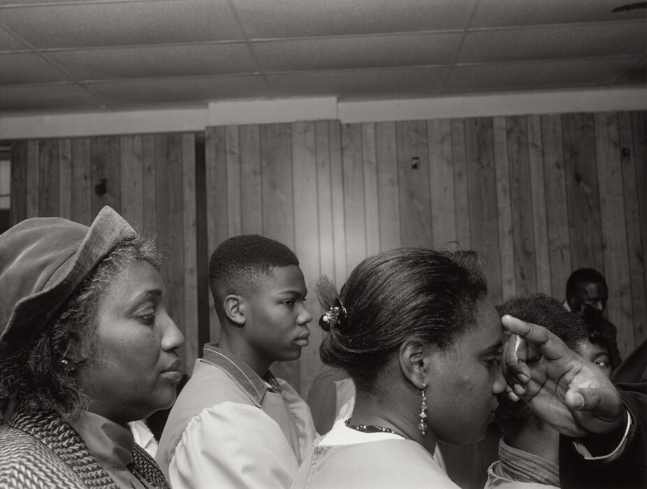 The image shows a partial view of several people's faces and upper bodies from the side. In the foreground on the left, an older woman is facing to the right, wearing a patterned coat and a hat. Beside her, a young man is visible in profile with an afro hairstyle, wearing a robe over a collared shirt. Next to him, a young woman is positioned facing slightly to the right, showing her profile and hair pulled back. She wears dangling earrings and possibly a necklace. Another person next to her has short hair. A hand is extended towards the person's face in the foreground. The background features a room with wood-paneled walls and a drop ceiling.