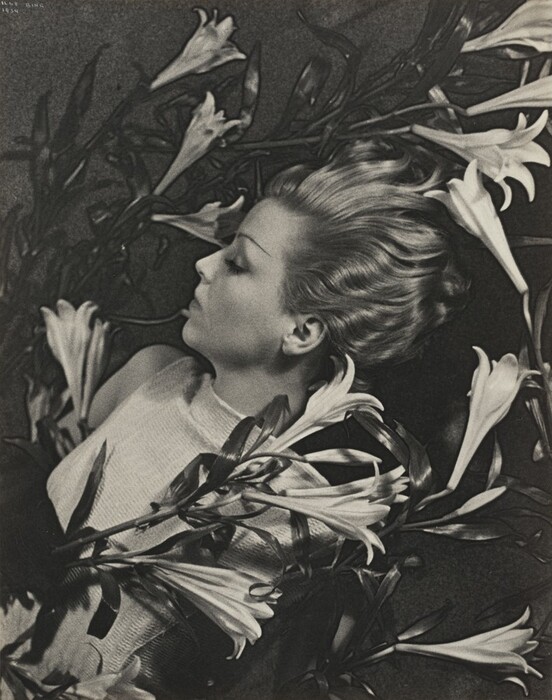 The image shows a woman reclining on a surface surrounded by lilies. She is in profile, facing left, with her eyes closed. The woman has a serene expression and styled hair, wearing a light-colored garment. The lilies frame her form and the background is dark. No jewelry is visible.