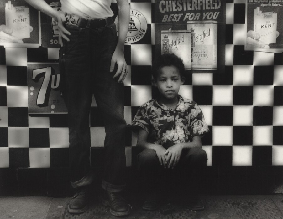 The image depicts two individuals standing against a checkered wall. On the left, only the lower half of a person is visible, wearing dark pants and a light-colored shirt. On the right, a young boy is sitting with his back against the wall, visible from head to mid-shin level. The boy has short curly hair and a neutral expression, wearing a patterned shirt and dark pants. The wall behind them is decorated with checkered tiles and advertising posters for cigarettes and 7 Up. The ground is visible at the bottom of the image, appearing as a concrete or similar surface.